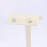 Load image into Gallery viewer, Gold bezel-set opal stud earrings on a cream display stand, showcasing iridescent blue and green hues.