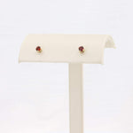 Load image into Gallery viewer, Gold stud earrings with deep red gemstones, elegantly displayed on a light beige stand. Perfect for any occasion.