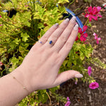 Load image into Gallery viewer, Delicate silver ring with blue gemstone, worn on hand among vibrant pink and green flowers in a outdoor setting.