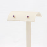 Load image into Gallery viewer, Gold stud earrings featuring round purple amethyst stones, elegantly displayed on a cream velvet stand.
