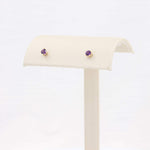 Load image into Gallery viewer, Gold stud earrings featuring round purple amethyst stones, elegantly displayed on a cream velvet stand.